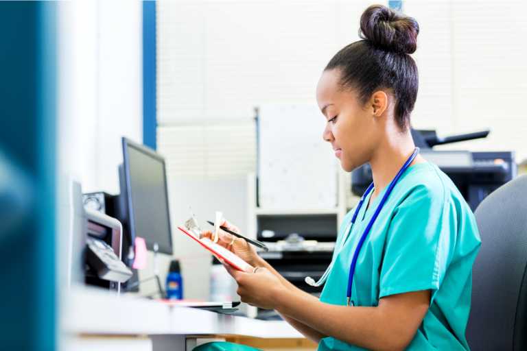 A healthcare professional in blue scrubs with a stethoscope around her neck is seated at a desk, intently reading a clipboard in a well-lit office with computer monitors in the background